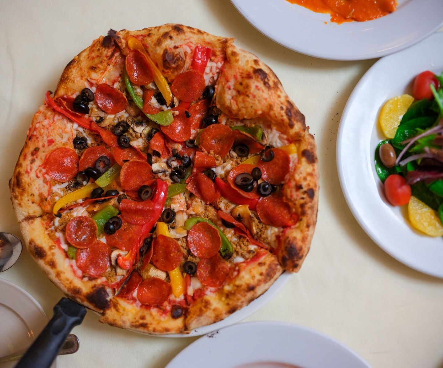 A pizza with pepperoni , olives , peppers and other vegetables is on a table with other plates of food.