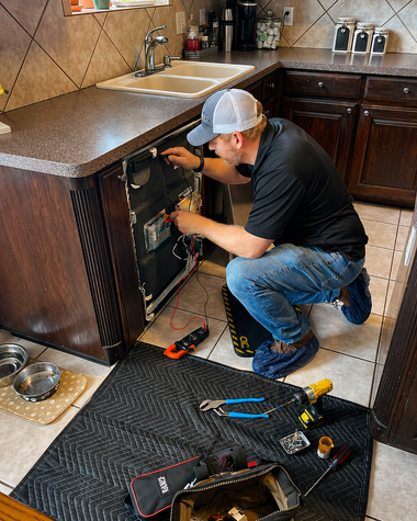 Technician kneels under kitchen counter, repairing an appliance with tools spread on the floor.