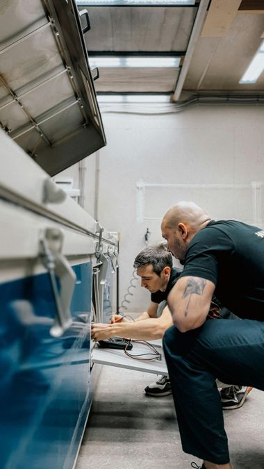 Two technicians crouch beside a blue machine, inspecting equipment in an industrial workshop.