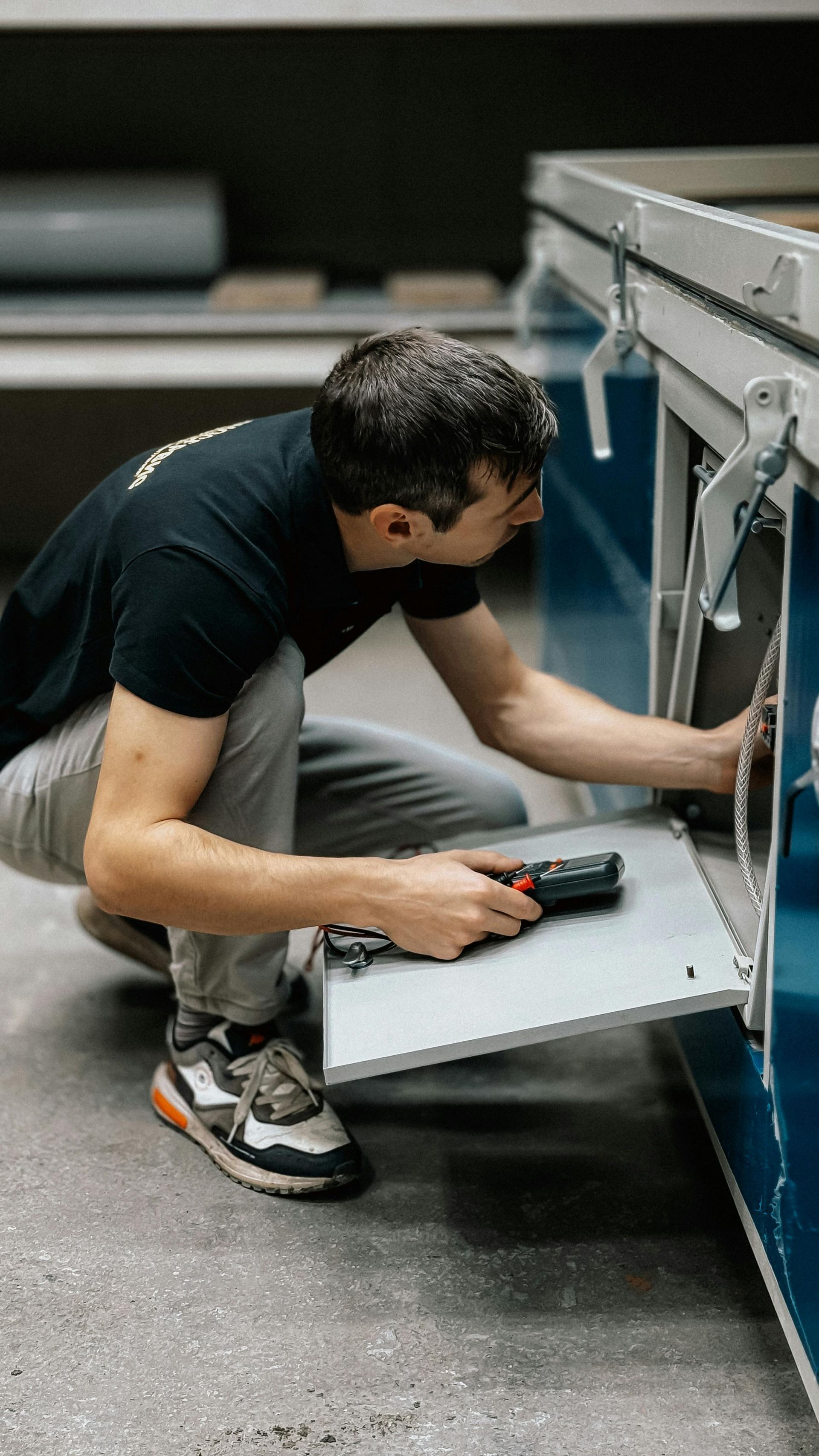 Person kneels beside a blue machine, using a tool to inspect an open panel.