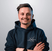 Man in a blue hoodie smiles, arms crossed, logo visible on hoodie, against a grey background.