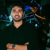 Man with a beard smiles, arms crossed. Dark clothing, watch visible. Dark background with blue lights.
