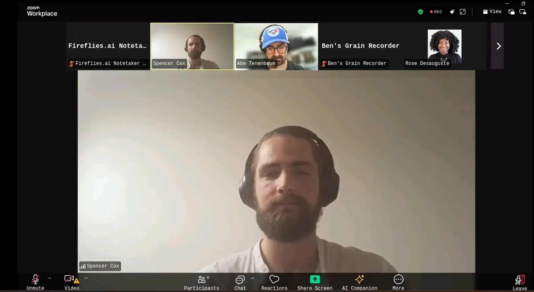 Video call with five participants. A man with headphones is the focus, with others visible in small boxes.