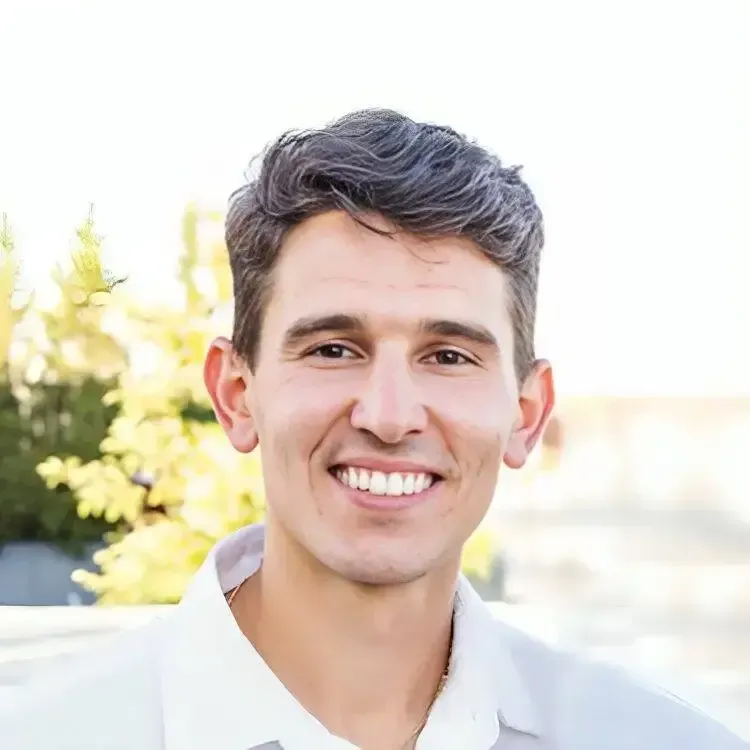 Man with dark hair smiling outdoors, wearing a white shirt; blurred greenery in the background.