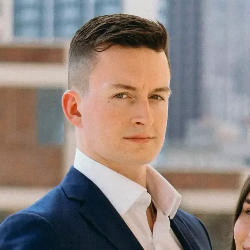 Man in a blue blazer and white shirt looking toward the camera; cityscape background.