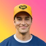 Man wearing a yellow cap with a black heart logo, smiling in front of a colorful background.