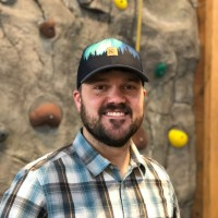Man smiling, wearing a white shirt and yellow sweater. Light gray background.