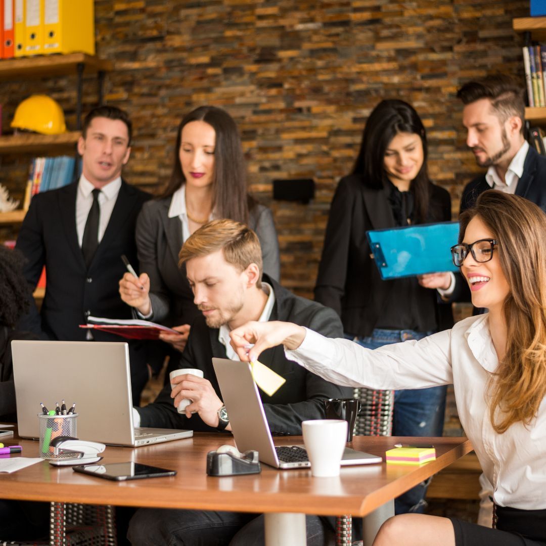 Group of people in an office. A woman points at something, while others look at laptops and papers.