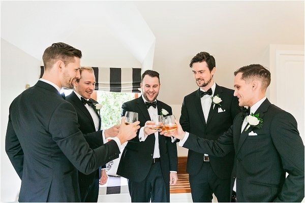 groom and 4 groomsmen having a drink and toast nefore leaving to the wedding. all in black with white buttonholes and distinguished