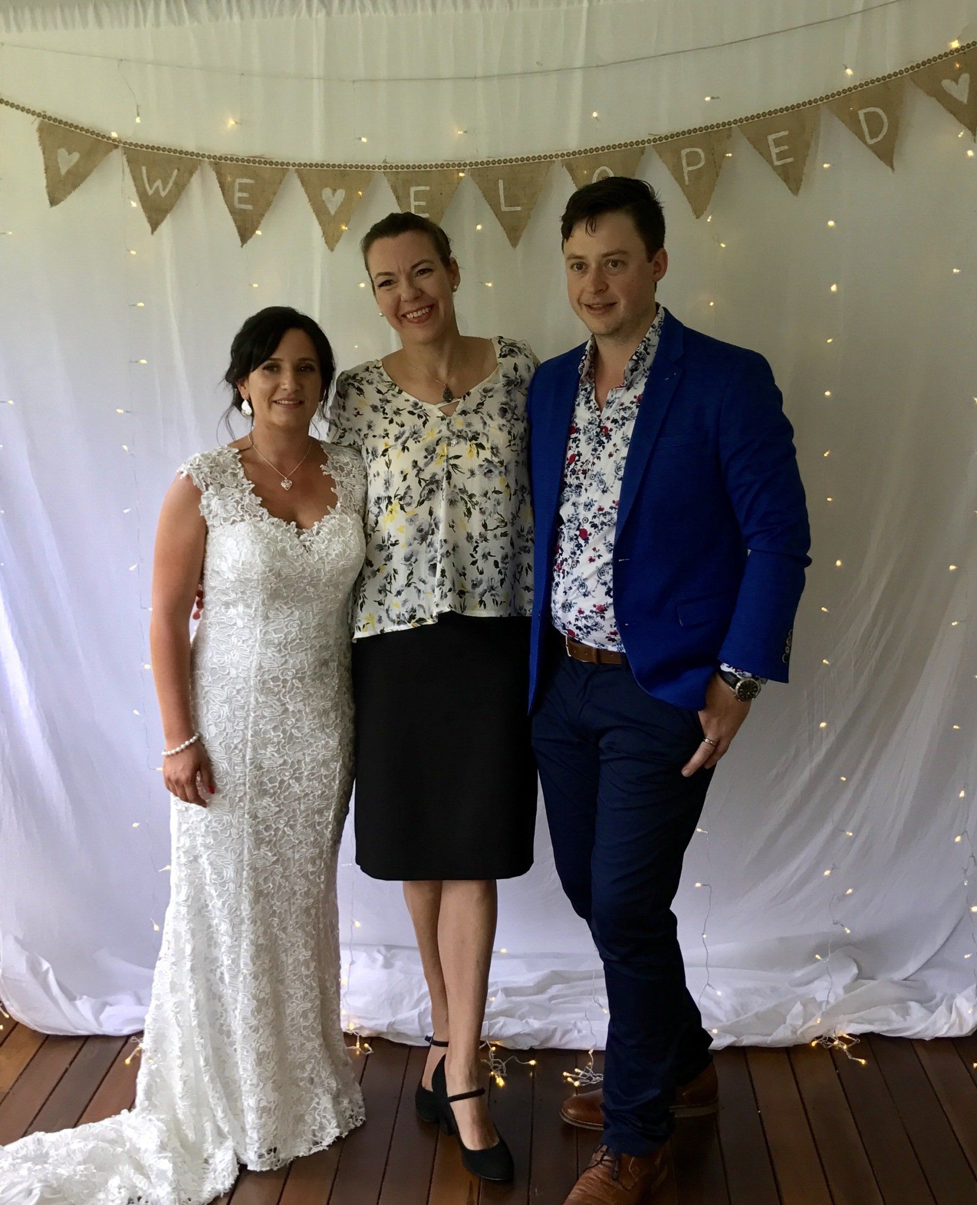 bridal couple elopement stand with celebrant under 'we eloped' sign and curtain with fairy lights at house