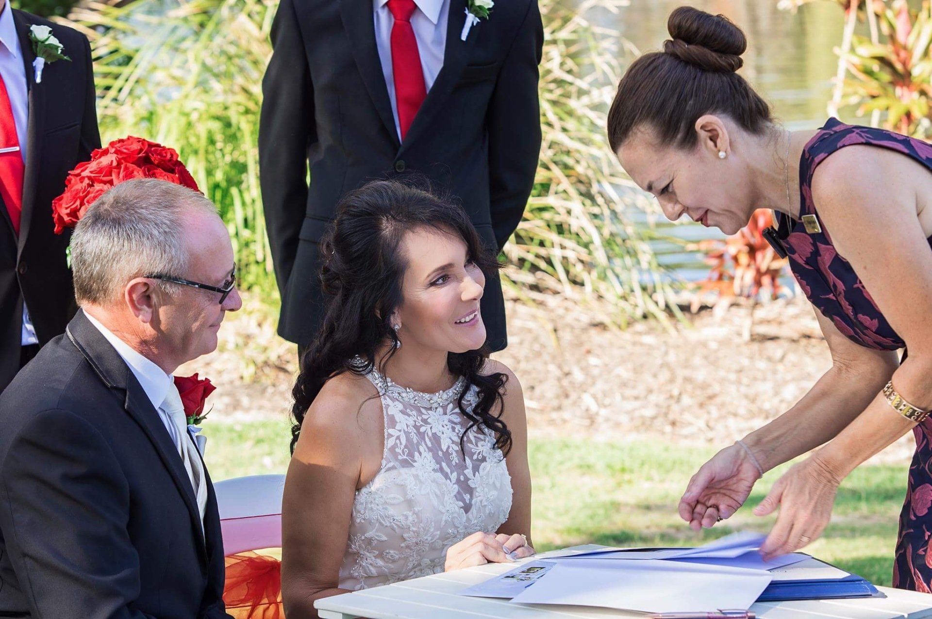 couple sit at signing table with red flowers after their Twin Waters Novotel wedding day