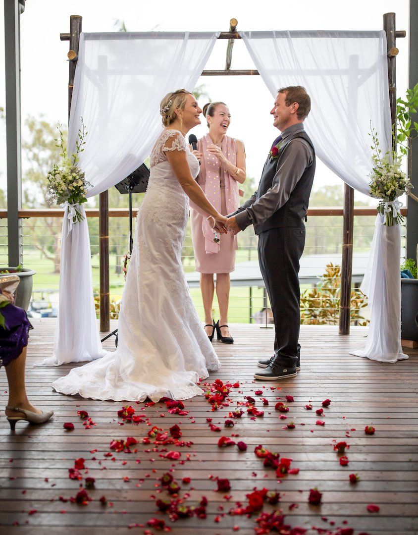 laughing couple under arbor with flowers being married by Noosa Celebrant in pink dress at Noosa Golf Club