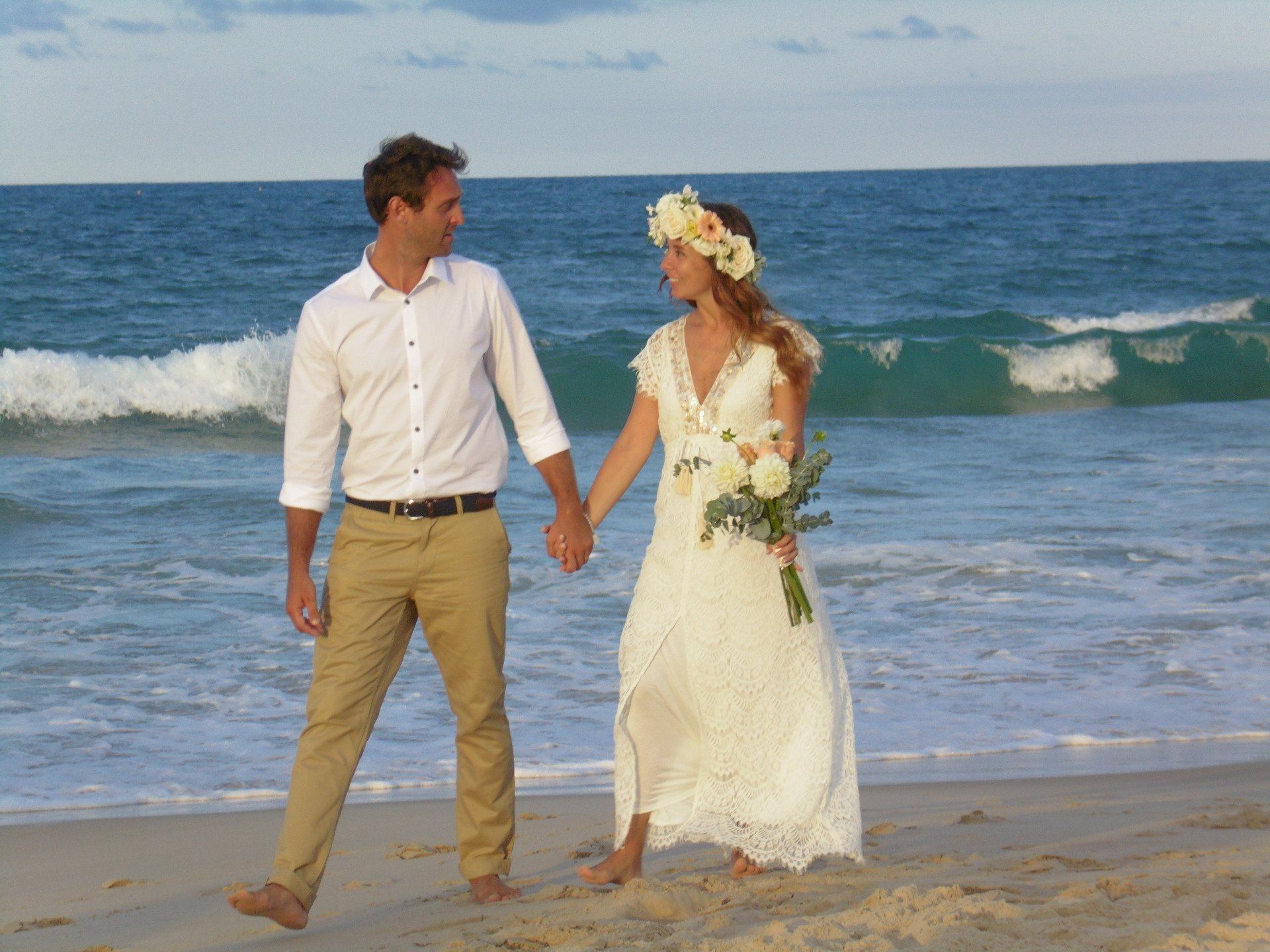 couple stroll in sand by ocean with flower bouquet hand in hand after their Noosa elopement