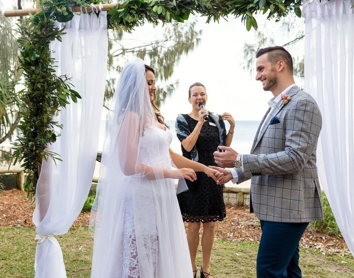 groom in checked jacket about to place wedding ring on wife's finger at Noosa wedding under white arbor
