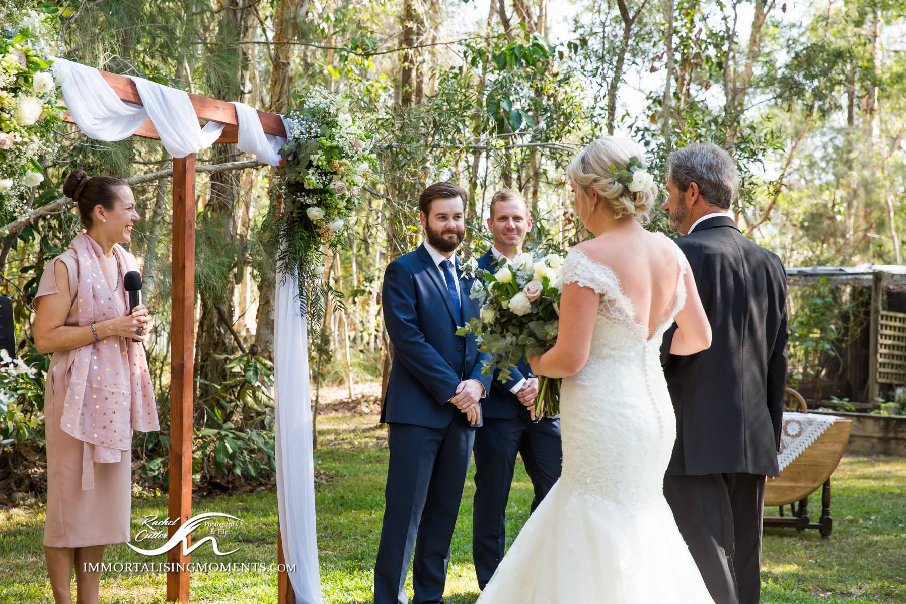 dad in grey suit walks his daughter down aisle to waiting groom in blue suit, smiling celebrant in pink ready to start wedding ceremony