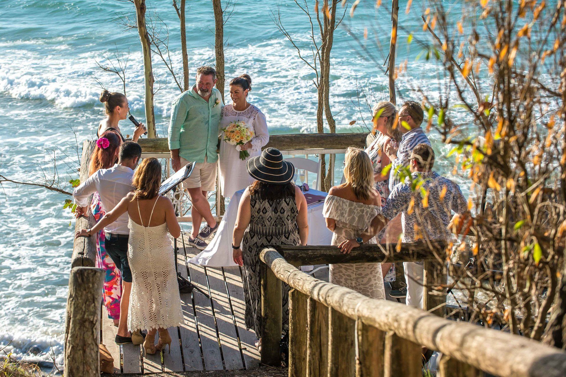 Noosa Little Cove platform Wedding bride holding yellow flowers in white dress, groom in green shirt, guests all happy