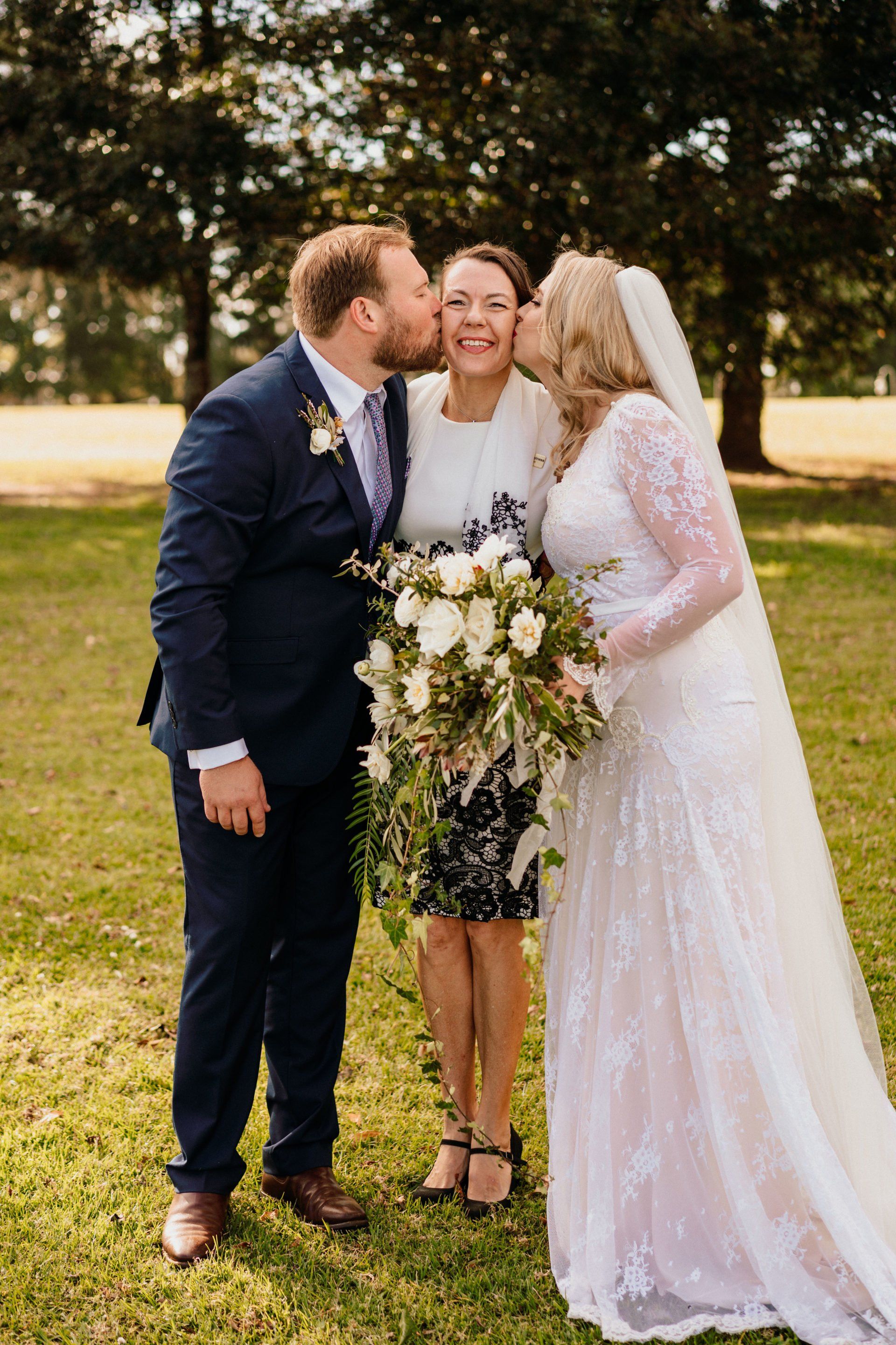 bride in white with veil and groom in blue suit kissing celebrant wearing black and white