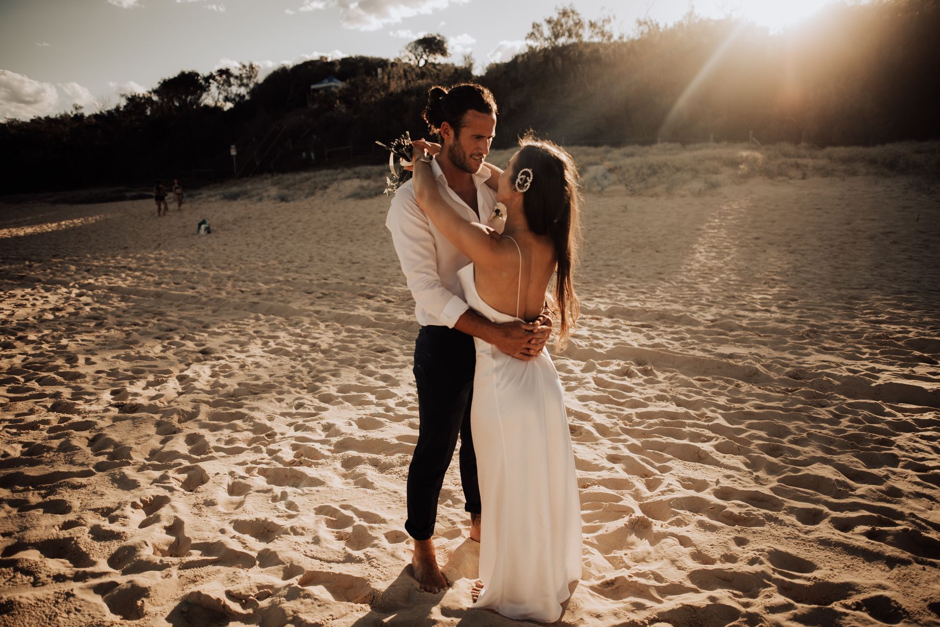 couple standing on Noosa Beach just married, embracing with sunset behind them, bride in white