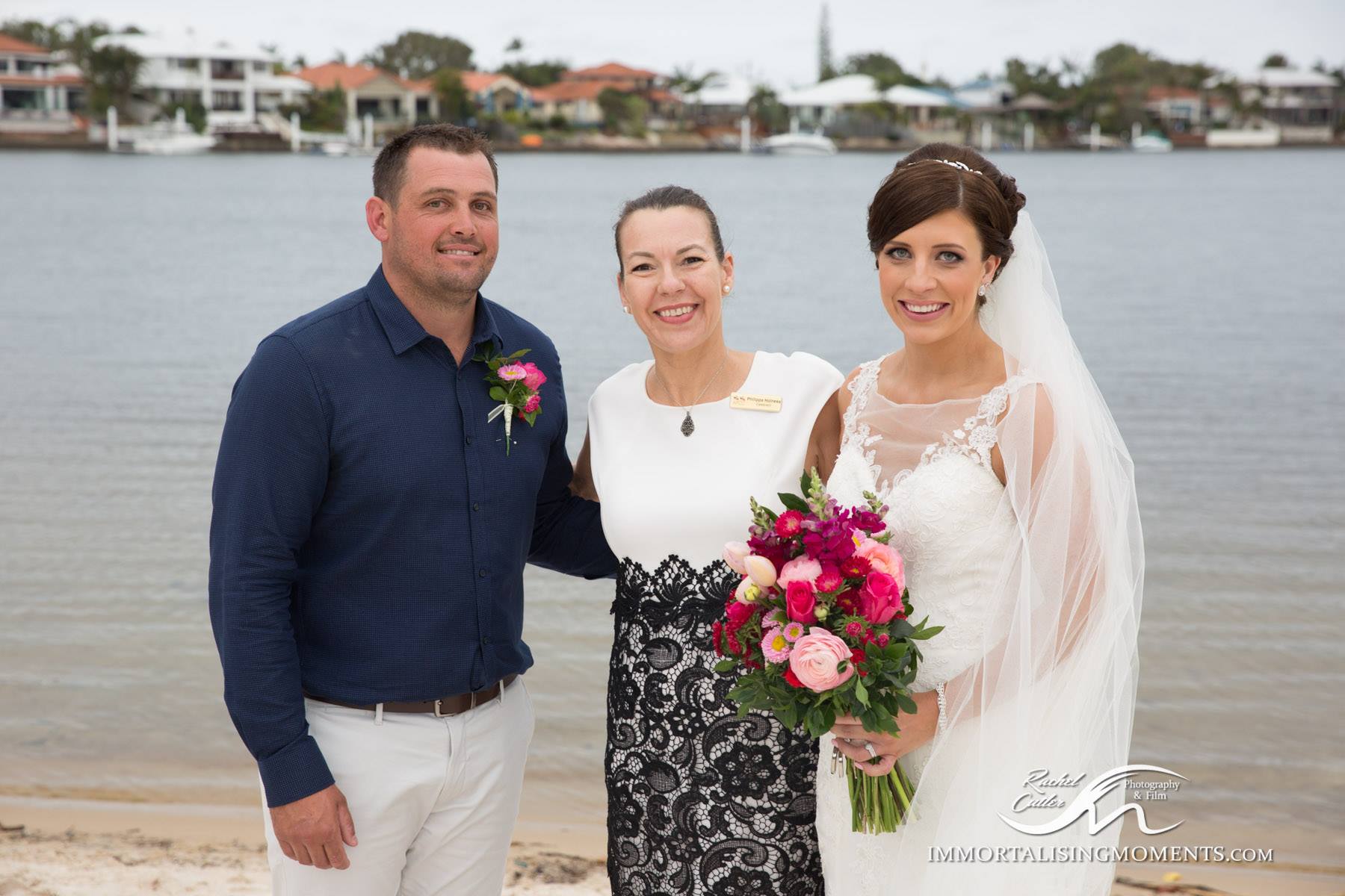 bride in white with pink bouquet and groom in blue shirt smiling with celebrant at Kawana Island by the water