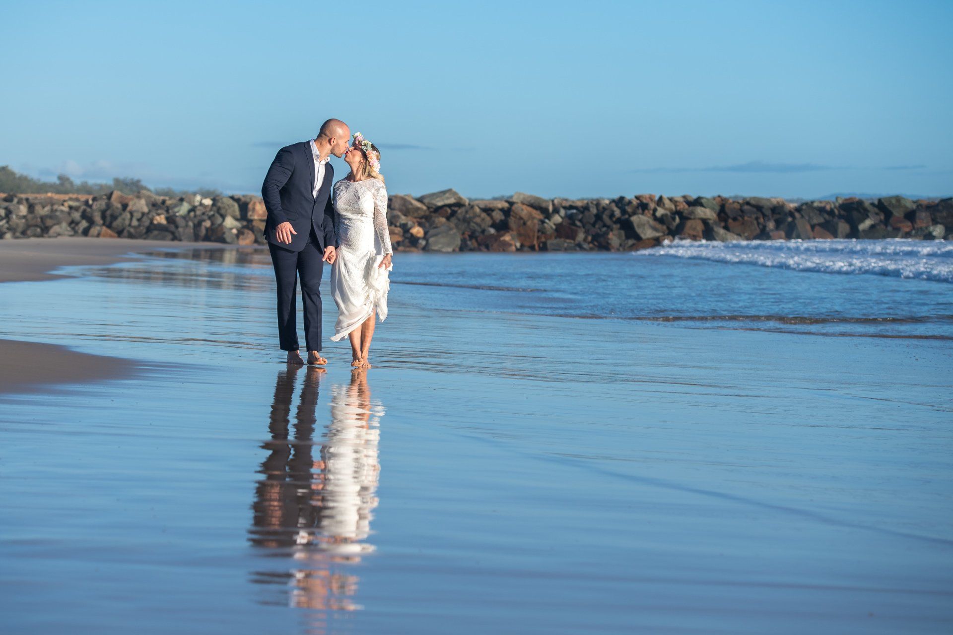 bride and groom kissing after their sunrise elopement in Noosa, walking in the ocean with their reflection visible by Noosa rockwall