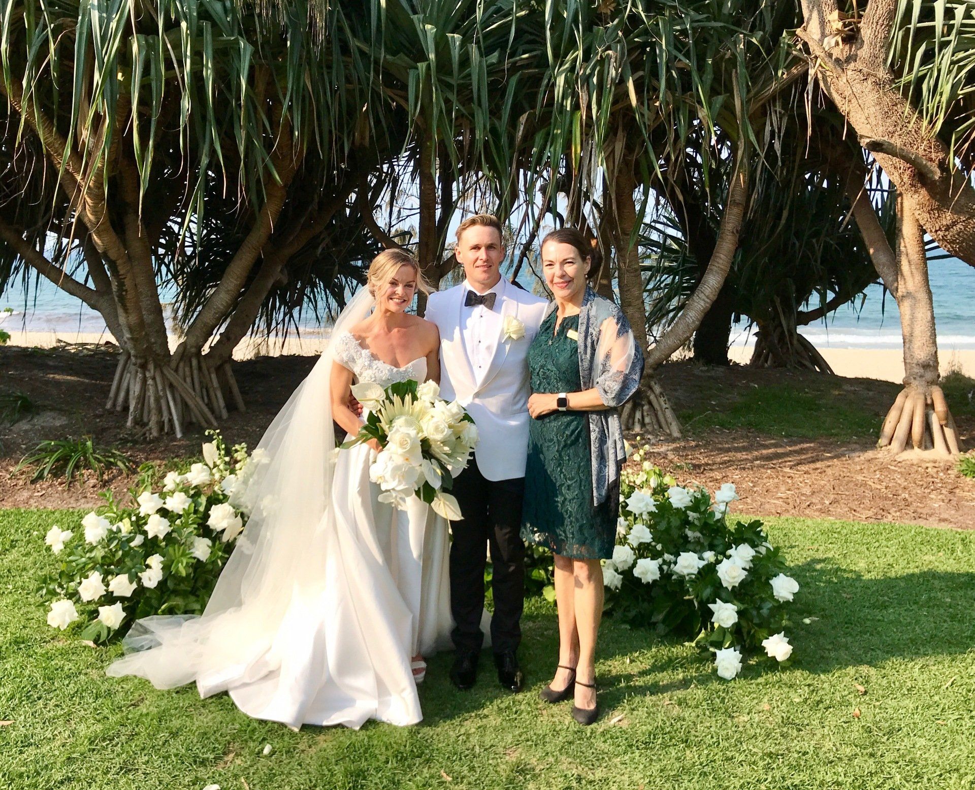 couple at maison la plage, Noosa with white and green floral installation standing with celebrant in green with sea behind them, bride with big white flowers