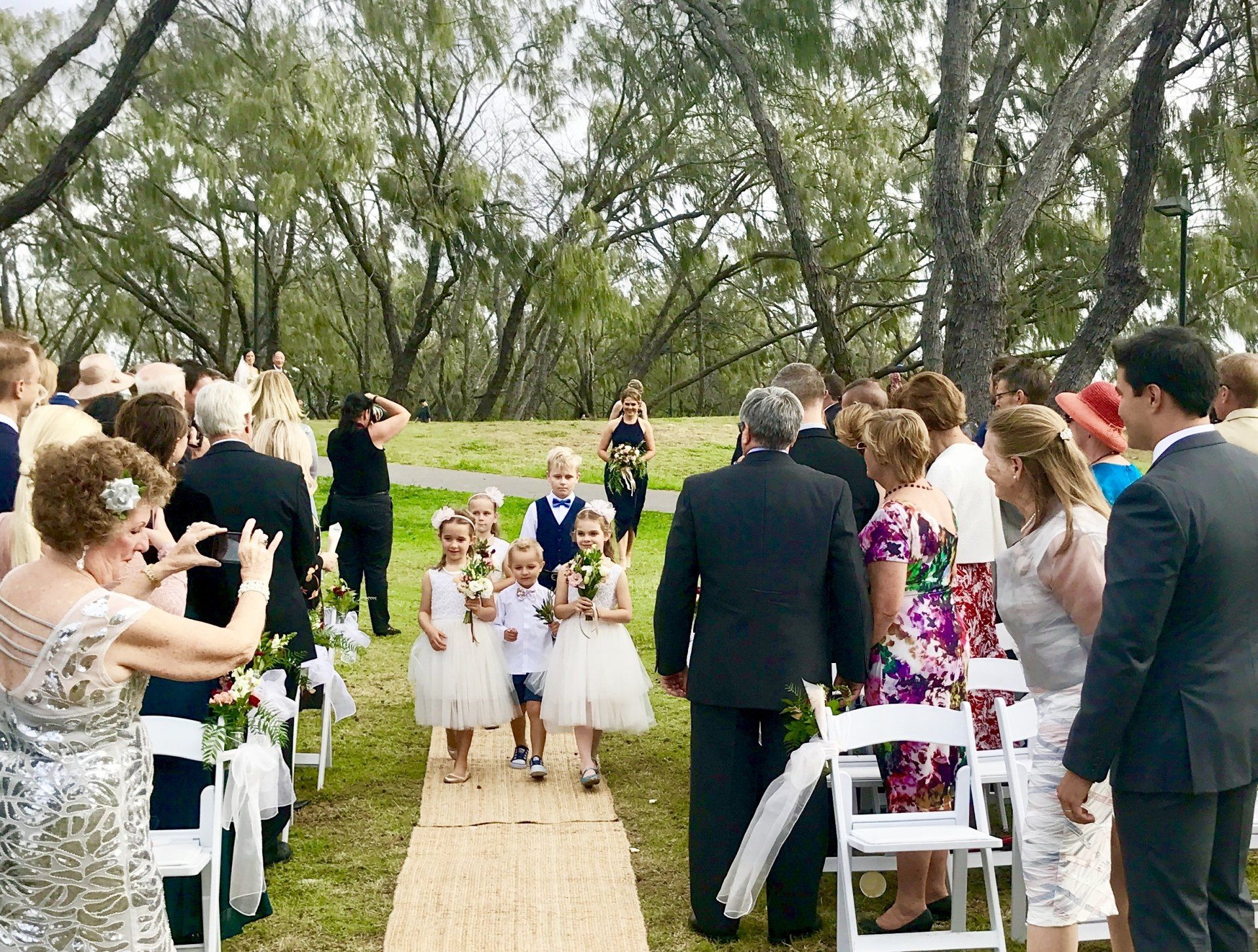 page boys and flower gilrs walk down the aisle with flowers before the bridesmaids in navy and the bride in white at Mooloolaba beach