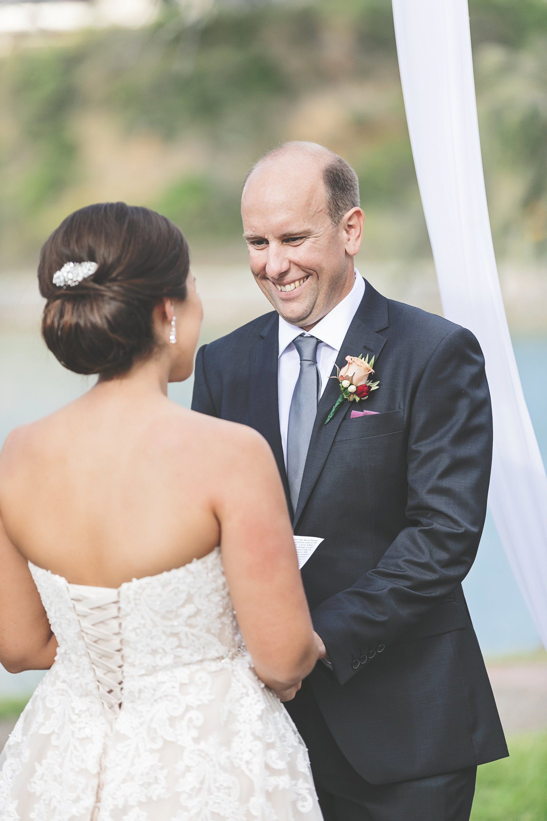 bride with dark brown elegant low bun reading her vows to bald happy husband in grey suit at their Mooloolaba beach wedding