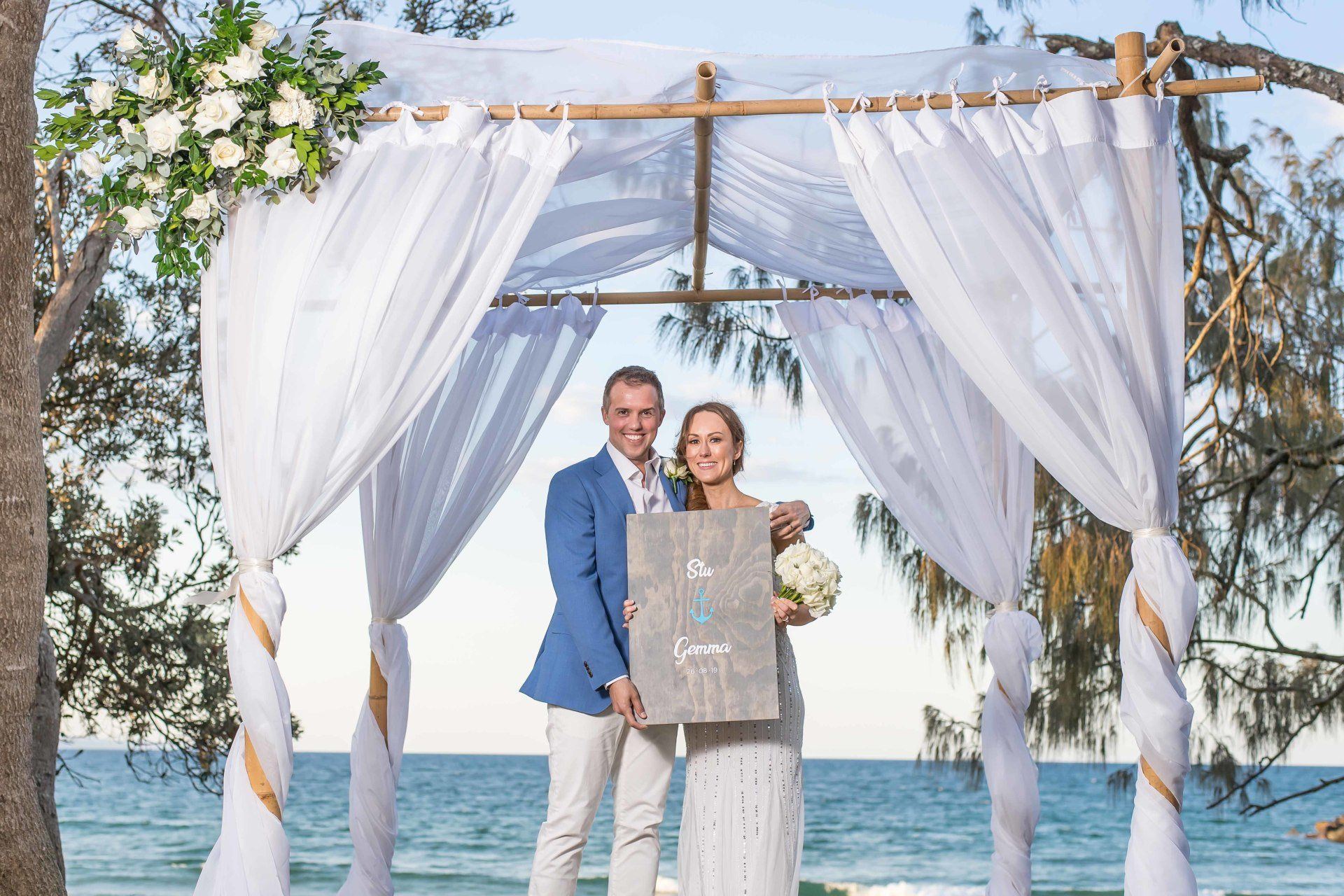 couple under wedding arbor holding sign with their names after their Wedding at Noosa beach Access 14/15