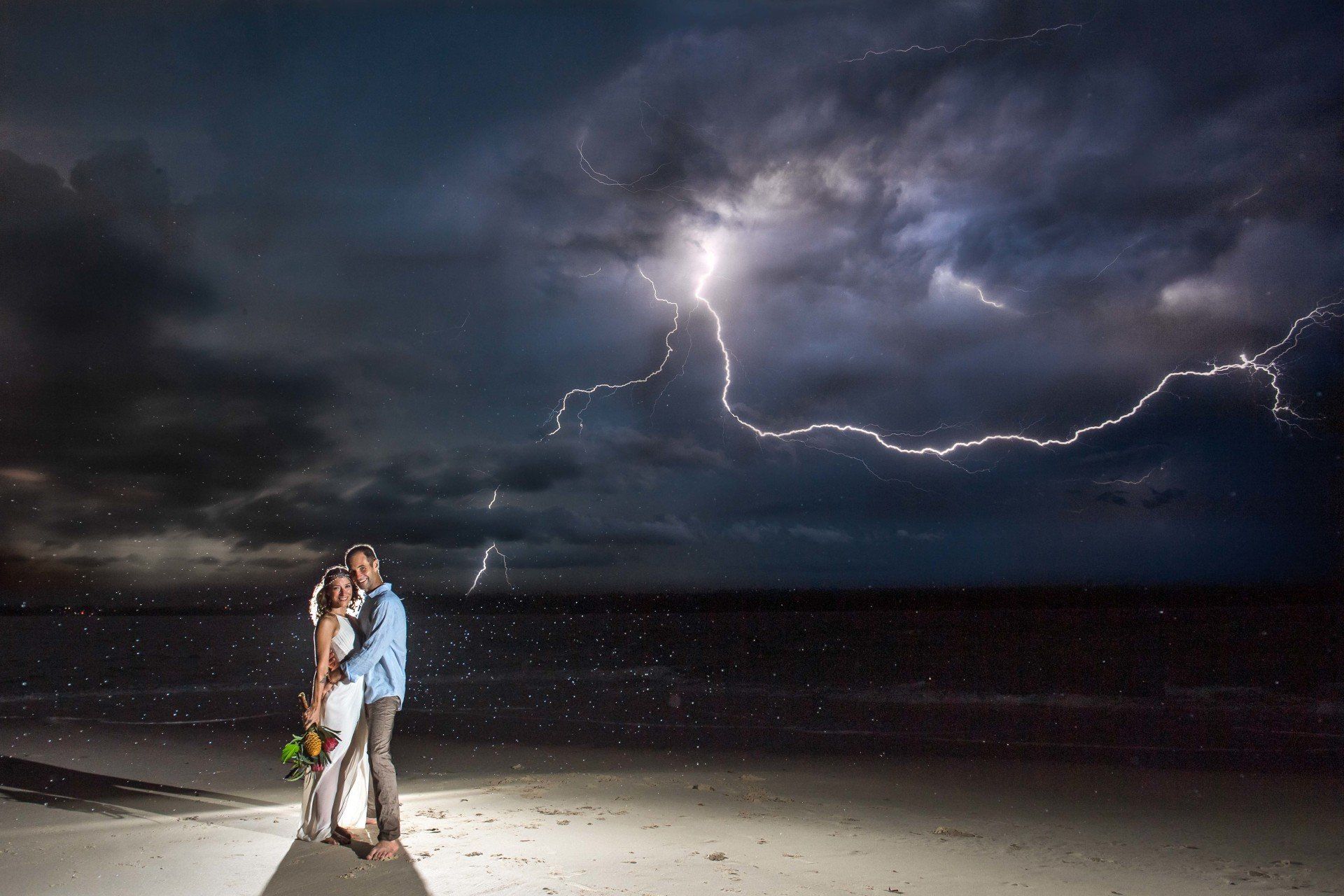 amazing lightening strike at night while just married couple are on beach embracing with flowers