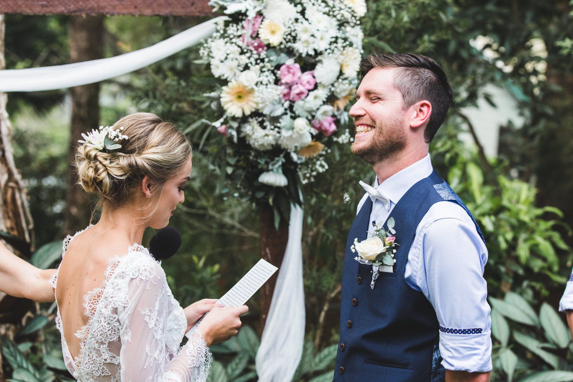 bride with flowers in hair and arbor with pink flowers reading her vows to her grinning husband wearing blue waistcoat