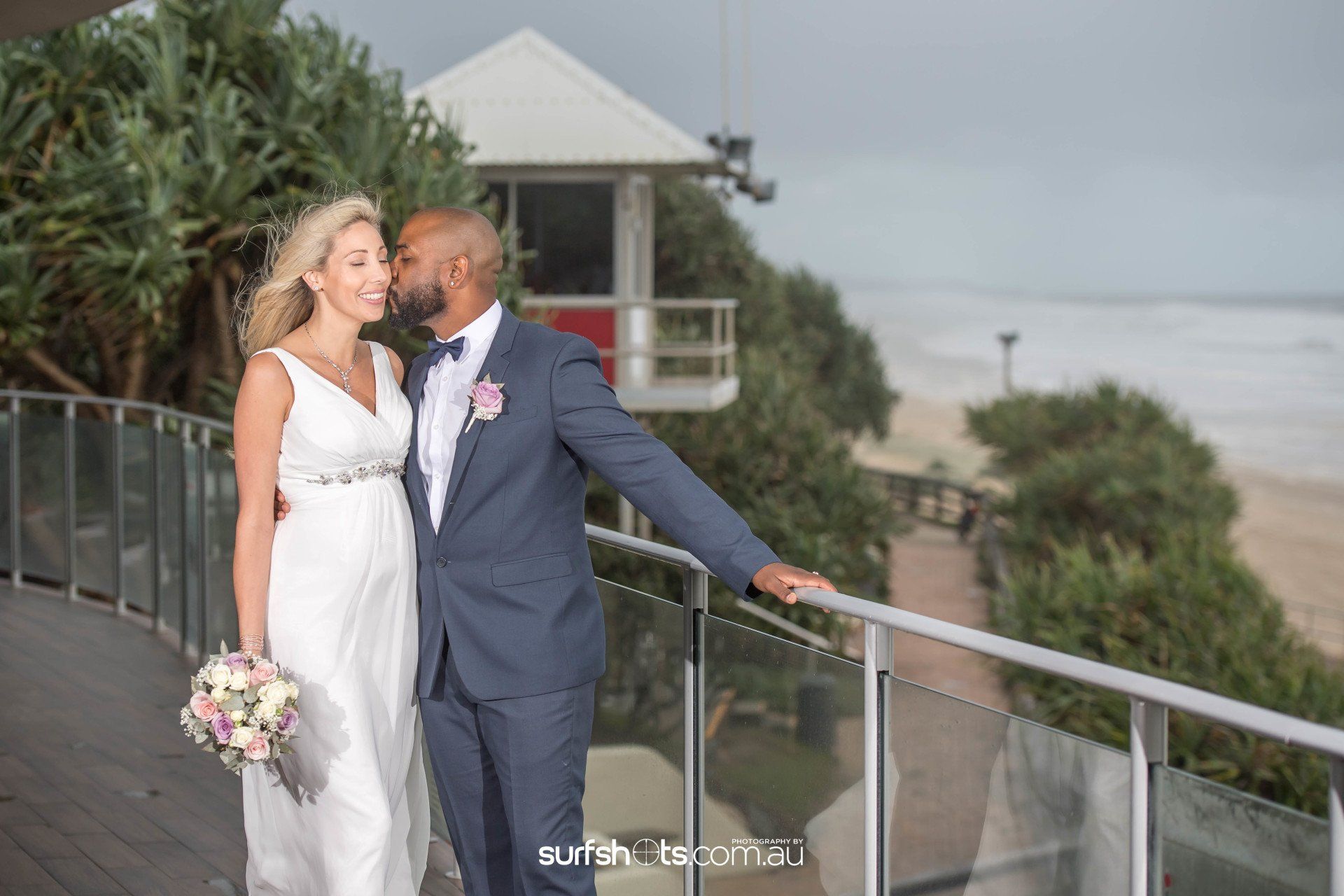 dark skinned groom kissing new wife on cheek after wedding, bride in white with pink flowers, on the deck at Coolum Surf Club with moody skies behind