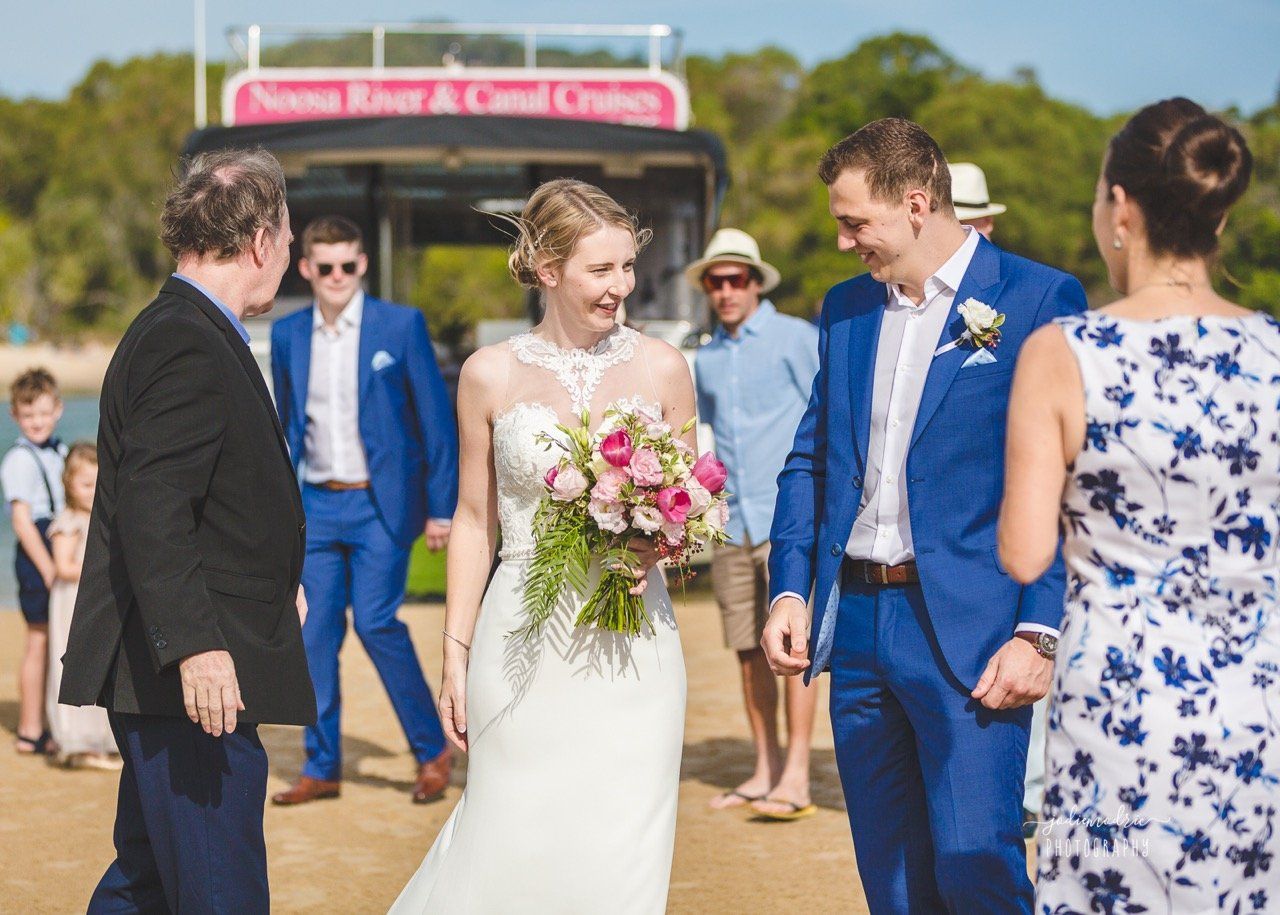 couple marrying on Noosa sandbank after cruising there in boat with their guests, groom in blue, bride has pink flowers