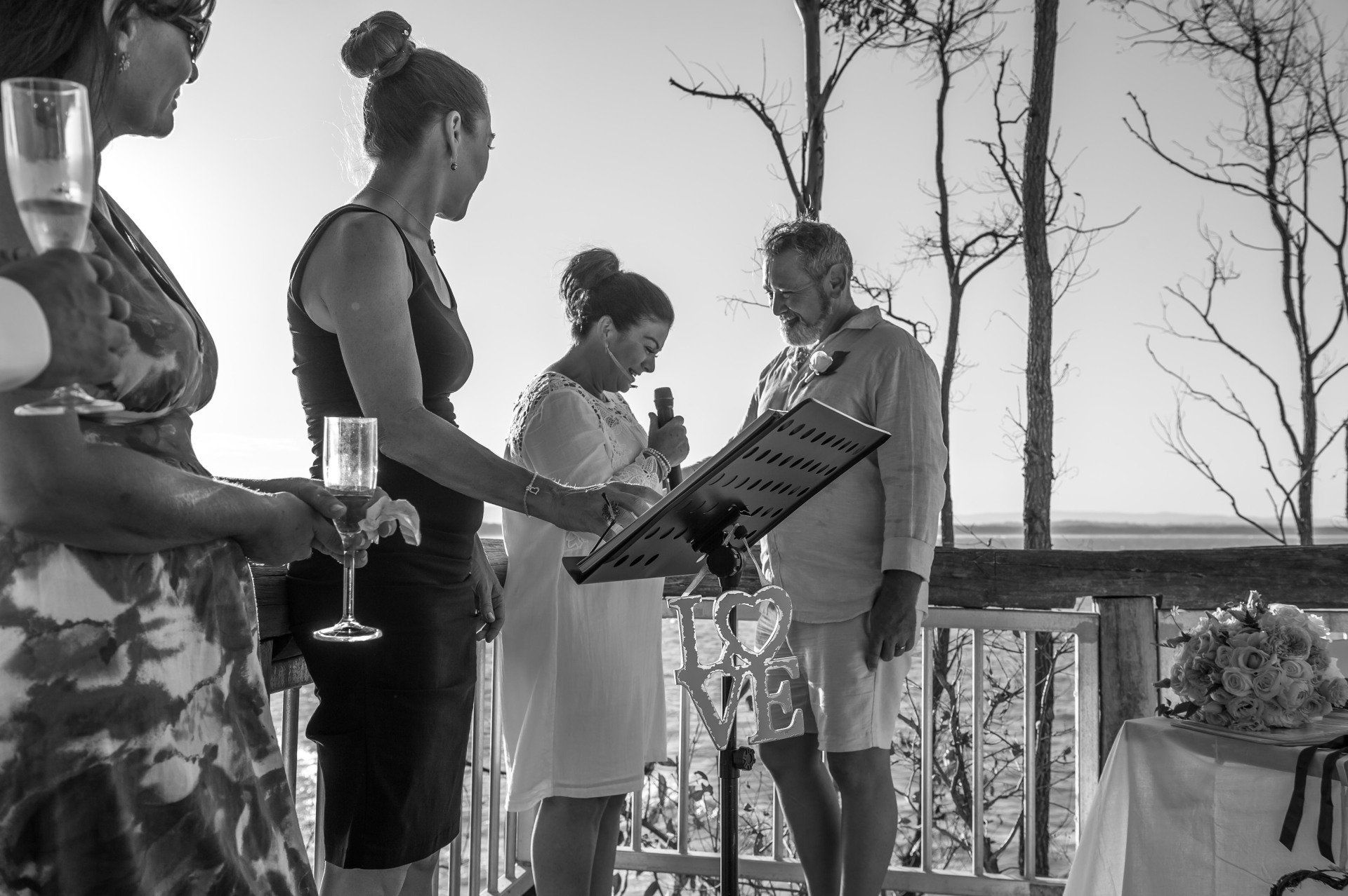 bride in white holding microphone saying her vows to her new husband, flowers on table and ocean behind at Little Cove deck in Noosa