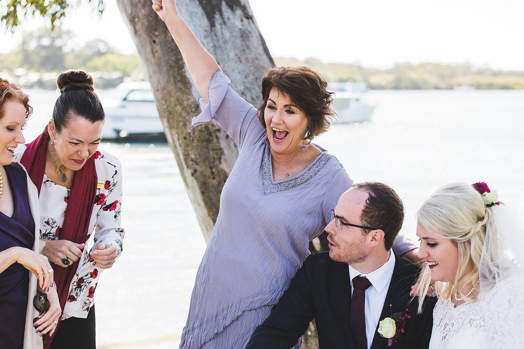 very excited mum wearing purple dress after being witness at her son's wedding, bride and groom sign paperwork at table, red themed wedding at Noosa River