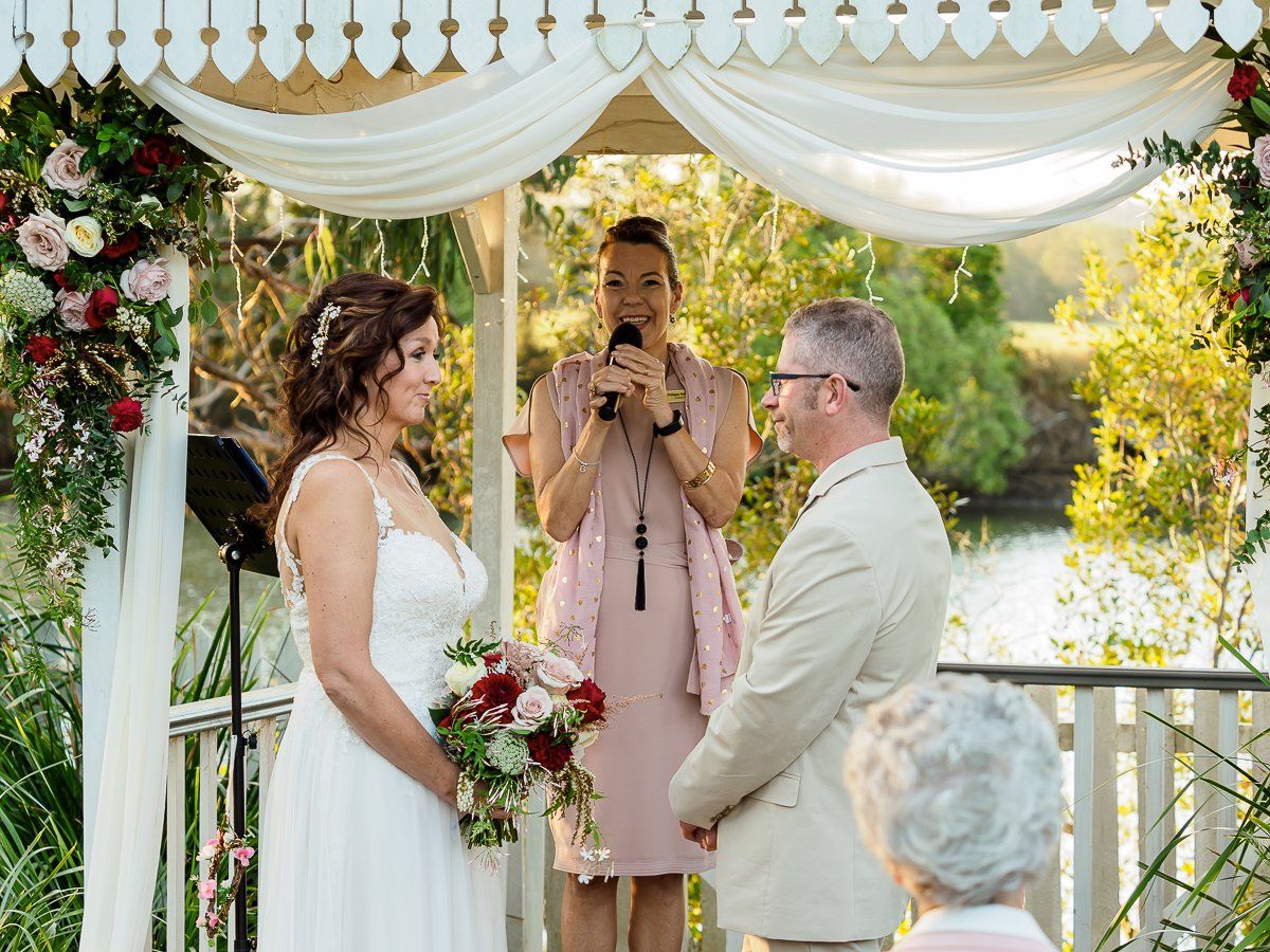 bride with red bouquet and archly hair clip marrying groom in beige suit, celebrant wearing pink telling their love story