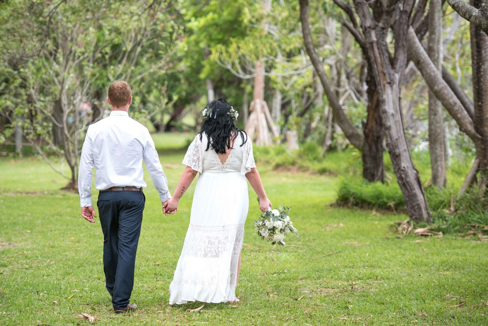 bride and groom stroll hand in hand through Noosa Woods after their Elopement , bride holding flowers in right hand