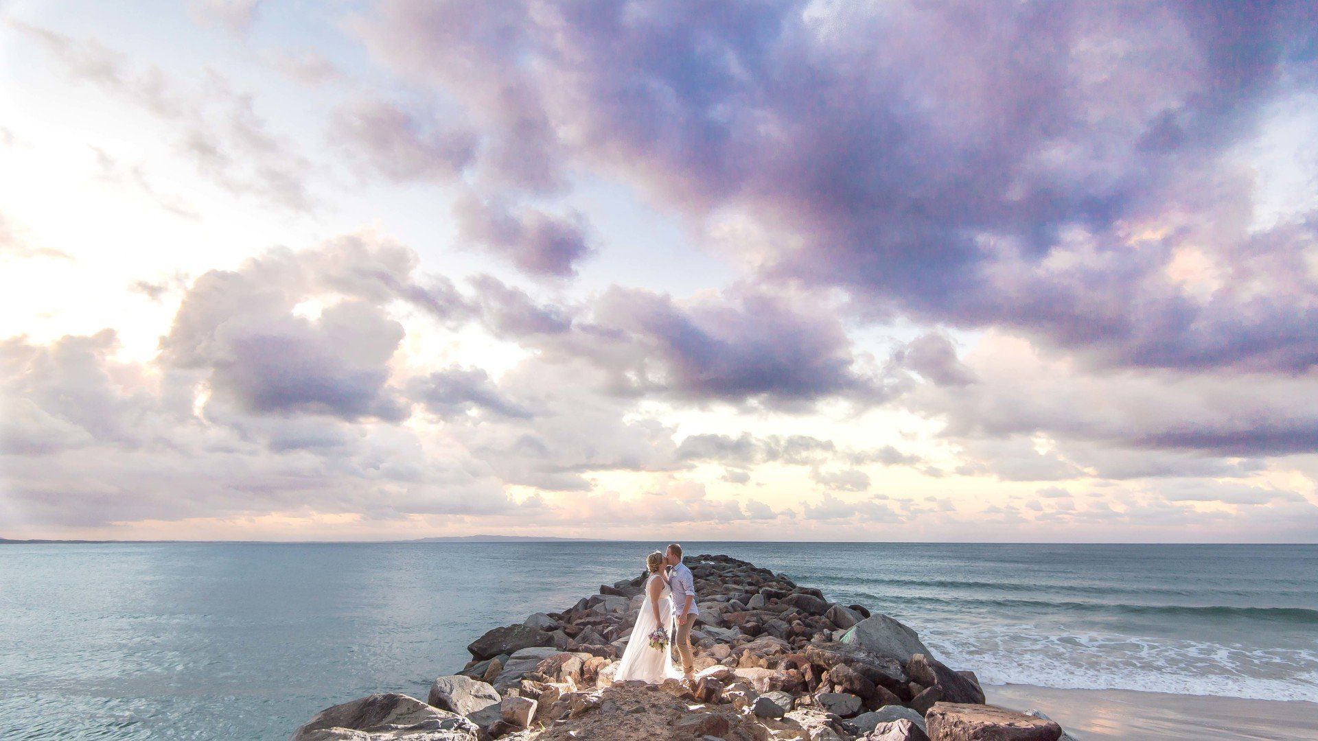 couple wearing white kiss on Noosa Rockwall at sunset after their elopement, with purple sky behind them