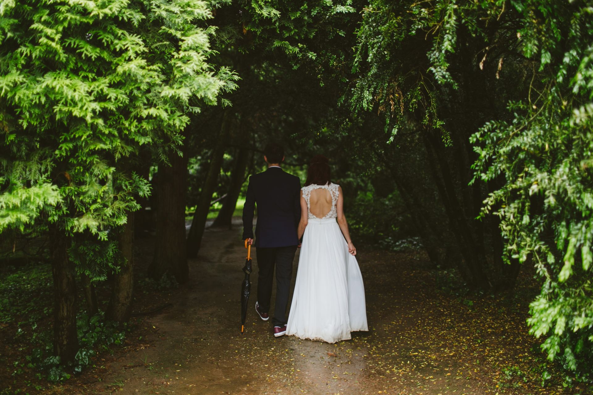 bridal couple walk through green tree covered park in wet weather with umbrella
