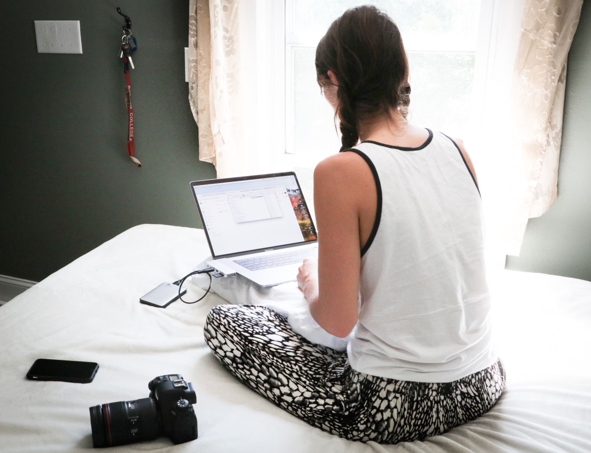 girl sitting on white bed with laptop on pillow and camera next to her searching on computer