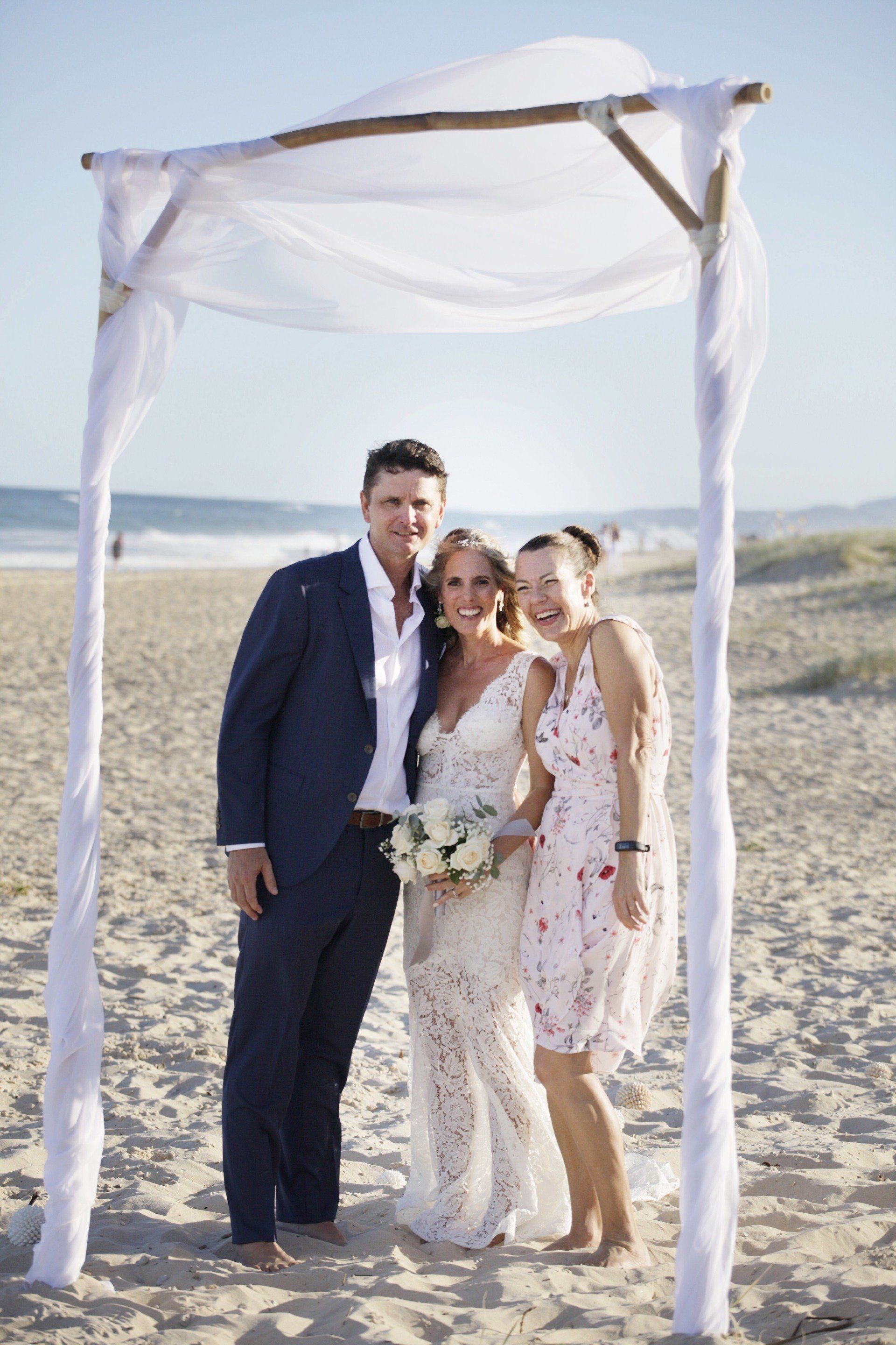 laughing bride and groom with Noosa celebrant in pink dress after the ceremony on Sunshine beach, beautiful white arbor and flowers