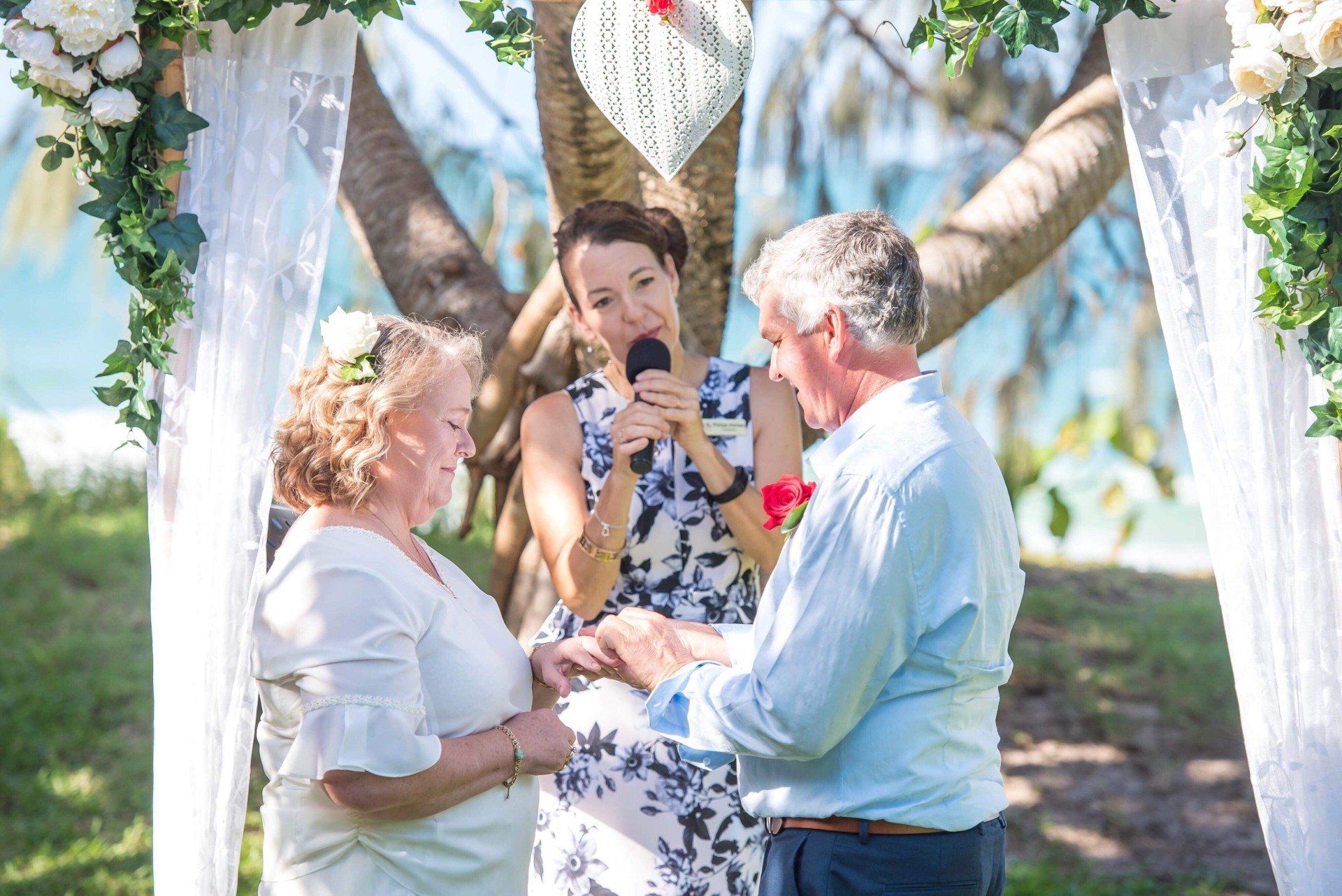 older couple putting rings on as the Noosa celebrant marries them under an arbor by the ocean