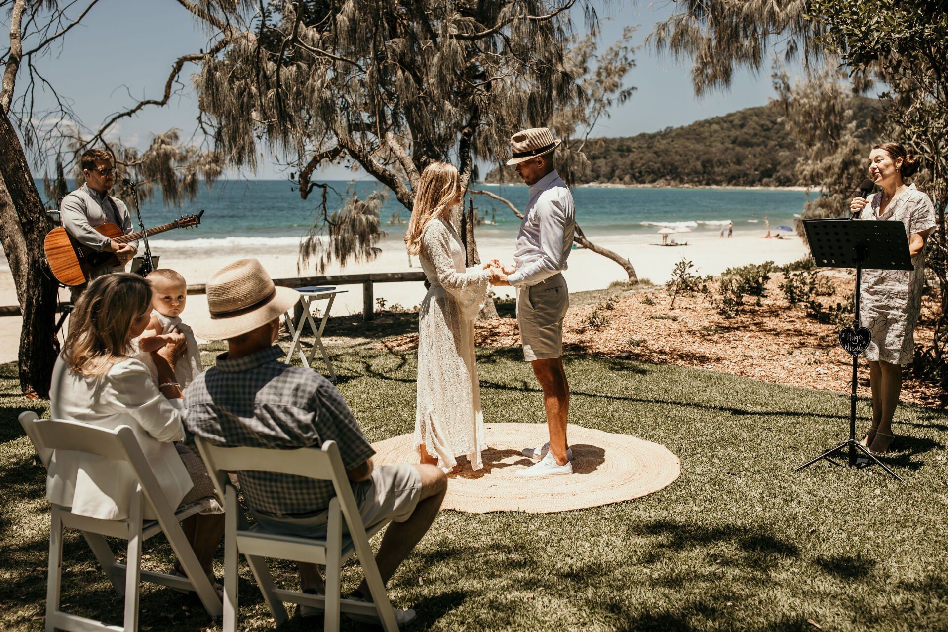 couple during a Noosa Elopement ceremony, with guest holding baby by the ocean at Hidden Grove, musician plays music for them