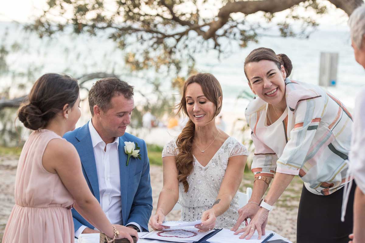 couple at atbel signing paperwork after their elopement in Noosa, ocean behind them and  Noosa celebrant pointing to where to sign
