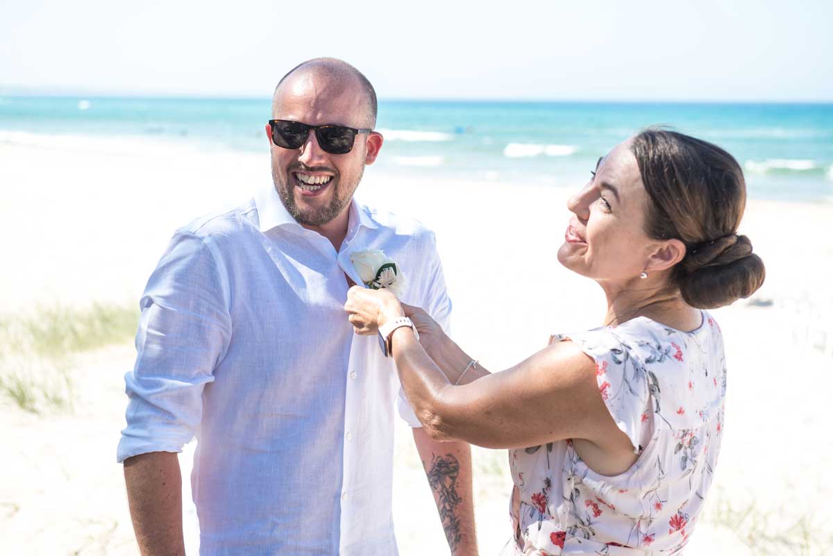 Noosa celebrant pinning white flower to groom's white shirt and smiling and chatting about the ceremony before bride arrives