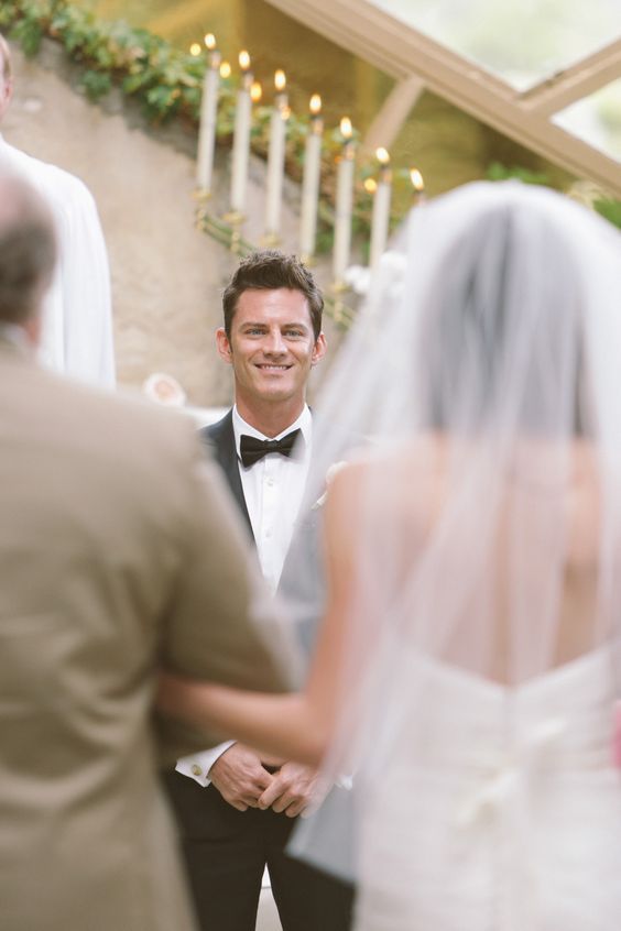 groom sees his bride in white veil walking with her dad for the first time, candles in background