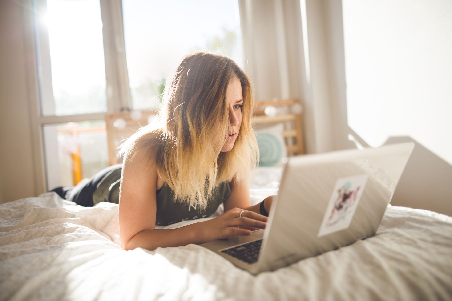 girl with straight blond hair and green singlet, laying on white bed looking at laptop