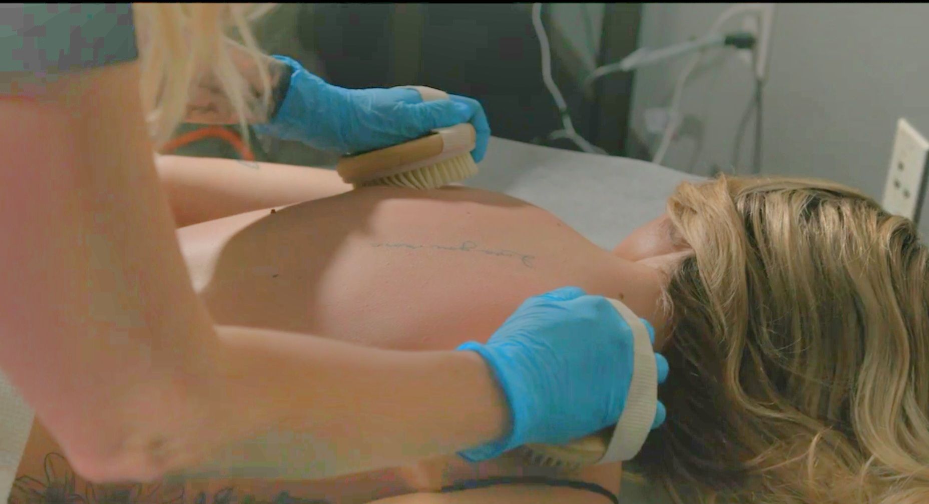 Two people brushing a person's back with blue gloves on, in a medical setting.