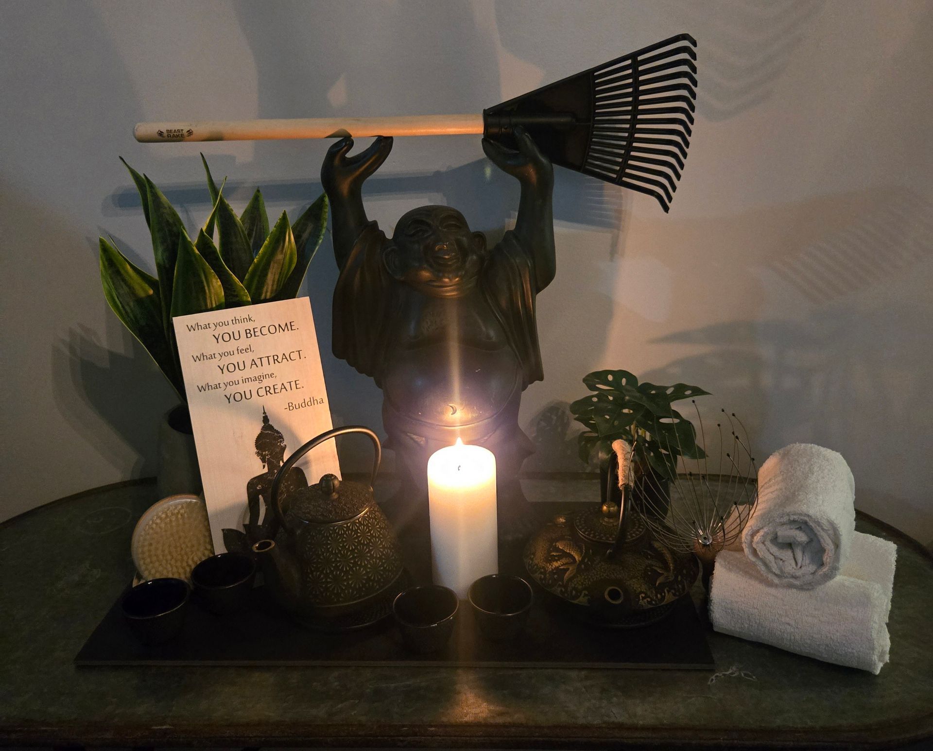 Zen-like altar: Buddha statue holding a rake, candlelit with plants, teapot, towels, and a book.