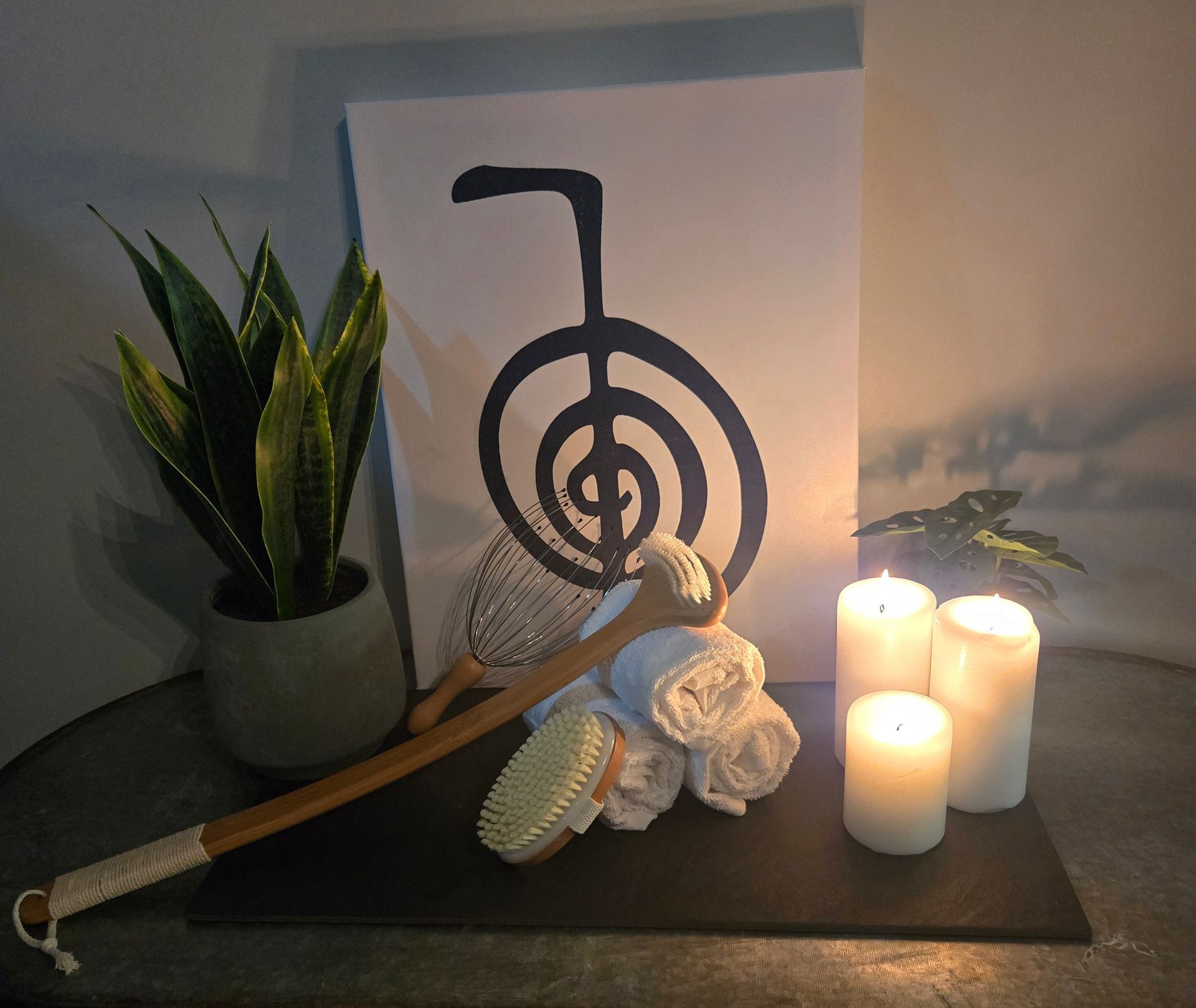 Relaxation scene: plant, Reiki symbol art, candles, towels, back brush on a dark surface.