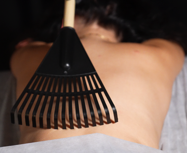Person receiving a massage with a rake-like tool on their back in a dimly lit room.